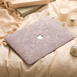 May include: A light lavender laptop cover featuring an embossed floral pattern with hibiscus flowers and leaves, with a central cutout revealing the silver Apple logo. The cover is placed on a soft, cream-coloured fabric surface.