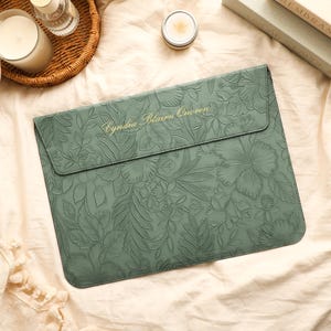 Puede incluir: Una funda para portátil de color verde salvia con un patrón floral en relieve en toda la superficie. La solapa incluye el texto dorado "Cyndia Blairn Quoren" en letra cursiva. La funda tiene forma rectangular con esquinas redondeadas.