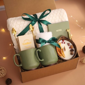 K&ouml;nnte beinhalten: Geschenkbox mit einer cremefarbenen Decke, zwei gr&uuml;nen Tassen mit Rillen, einer Kerze und einem Notizbuch. Goldene L&ouml;ffel und ein gr&uuml;nes Band schm&uuml;cken die Box. Die Kerze befindet sich in einer Kokosnussschale.