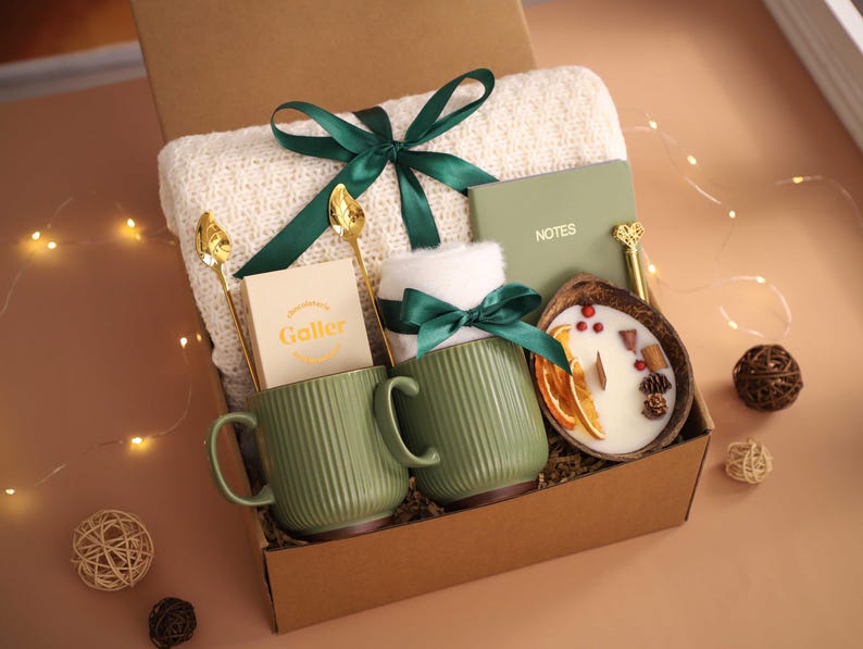 K&ouml;nnte beinhalten: Geschenkbox mit einer cremefarbenen Decke, zwei gr&uuml;nen Tassen mit Rillen, einer Kerze und einem Notizbuch. Goldene L&ouml;ffel und ein gr&uuml;nes Band schm&uuml;cken die Box. Die Kerze befindet sich in einer Kokosnussschale.