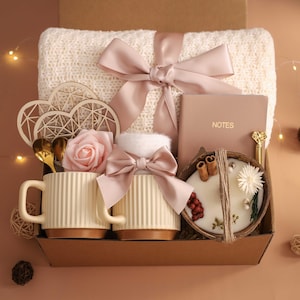 K&ouml;nnte beinhalten: Eine Geschenkbox gef&uuml;llt mit einer cremefarbenen Decke, zwei Tassen, einem Notizbuch, einer Kerze und dekorativen Elementen. Die Tassen sind cremefarben mit braunen B&ouml;den. Eine rosa Schleife schm&uuml;ckt die Decke. Die Box enth&auml;lt eine Rose, Holzherzen und goldene L&ouml;ffel.