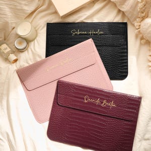 May include: Three rectangular laptop sleeves with a textured crocodile skin pattern in black, light pink, and deep burgundy. Each sleeve features a flap closure and gold cursive text on the front, reading Sabrina Harlen, and Querida Benton.