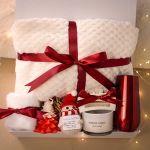 Può includere: Un set regalo con una coperta strutturata color crema, legata con un nastro bordeaux. Il set include un bicchiere rosso, una candela profumata, un ornamento a forma di pupazzo di neve e un piccolo oggetto bianco e soffice legato con un nastro bordeaux. Gli articoli sono disposti in una scatola bianca.