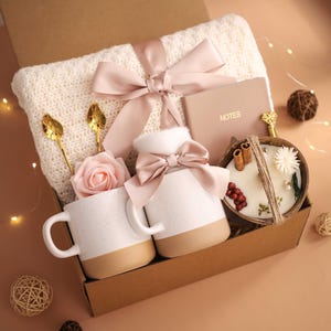 K&ouml;nnte beinhalten: Eine Geschenkbox mit einer cremefarbenen Strickdecke, zwei wei&szlig;en Tassen mit beigen B&ouml;den, einer Kerze in einer Kokosnussschale, einem Notizbuch und dekorativen Elementen. Die Box ist mit einem rosa Band gebunden und enth&auml;lt goldene L&ouml;ffel und eine rosa Rose.