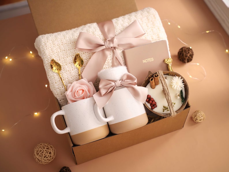 K&ouml;nnte beinhalten: Eine Geschenkbox mit einer cremefarbenen Strickdecke, zwei wei&szlig;en Tassen mit beigen B&ouml;den, einer Kerze in einer Kokosnussschale, einem Notizbuch und dekorativen Elementen. Die Box ist mit einem rosa Band gebunden und enth&auml;lt goldene L&ouml;ffel und eine rosa Rose.