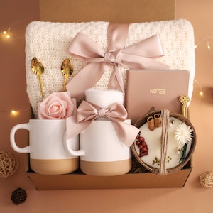 K&ouml;nnte beinhalten: Eine Geschenkbox mit einer cremefarbenen Decke, zwei wei&szlig;en Tassen mit beigen B&ouml;den, einer rosa Rose, einem Notizbuch mit "NOTES", einer Kerze in einer Kokosnussschale und goldenen L&ouml;ffeln. Die Box ist mit einem rosa Band gebunden.