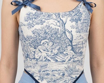 May include: A white and blue toile print corset top with a landscape scene. The top has blue ribbon straps and trim. The corset is paired with light blue pants.