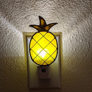 Op de afbeelding: Een geel ananasvormig nachtlampje dat in een wit stopcontact is gestoken. Het licht is aan en verlicht de ananas.
