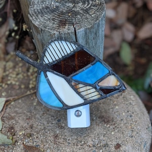 May include: A stained glass night light shaped like a butterfly with blue, brown, white, and beige glass pieces. The night light is plugged into an electrical outlet.