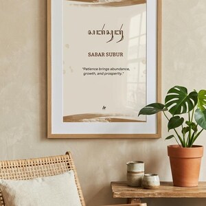 Op de afbeelding: Een ingelijste wandkunstprint met een lichtbeige achtergrond en bruine penseelstreekaccenten aan de boven- en onderkant. De tekst toont Sabar Subur in Javaans schrift, gevolgd door de woorden Sabar Subur en het citaat Patience brings abundance, growth, and prosperity.