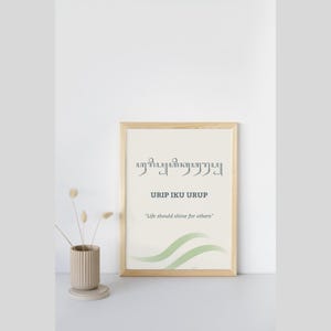 Può includere: Una stampa artistica da parete incorniciata con caratteri giavanesi, il testo "URIP IKU URUP" e la traduzione inglese "Life should shine for others" in grigio scuro. Il design include grafiche a onde verde tenue su sfondo color crema.