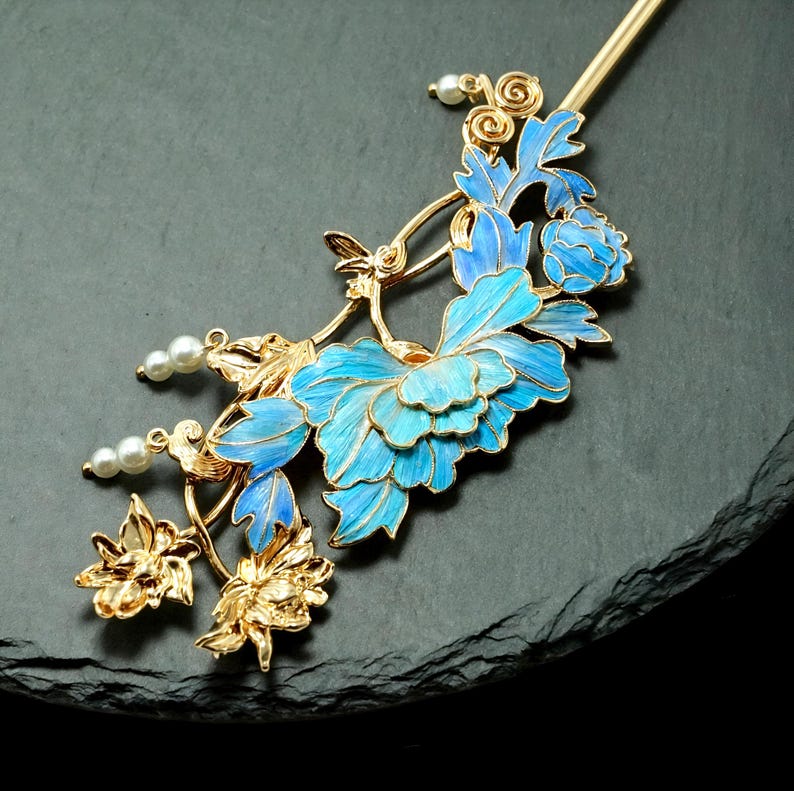 Pode incluir: Um gancho de cabelo dourado com um design floral intrincado com p&eacute;talas e folhas em esmalte azul vibrante. O acess&oacute;rio &eacute; acentuado com pequenas contas brancas redondas e detalhes delicados em filigrana dourada, criando uma apar&ecirc;ncia bot&acirc;nica.