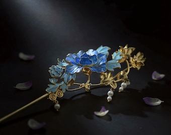 May include: A gold-toned hair pin featuring a blue floral design with light blue leaves and small white pearls. The flower has a detailed enamel finish. The pin is set against a dark background with scattered petals.