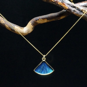 May include: A gold-colored necklace with a pendant featuring a blue, fan-shaped design. The pendant has a gold-colored border and a textured, iridescent blue center. The necklace hangs against a dark background.