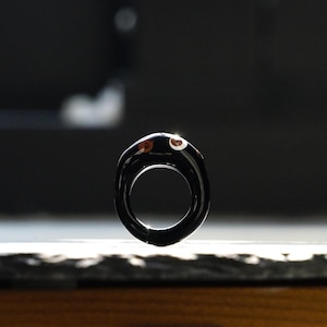May include: A black ring with a distinctive, flowing form. The ring has a small decorative detail with brown and white patterns. The design is contemporary and minimalist, suitable for everyday wear or special events.