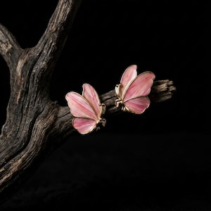 Puede incluir: Un par de pendientes con forma de mariposa con alas rosas y blancas y ribetes dorados. Los pendientes se exhiben sobre una rama oscura y texturizada sobre un fondo negro. Joyas.
