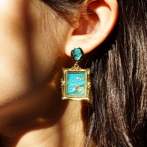 May include: A gold-toned earring featuring a rectangular frame with an ornate border. The center displays a textured blue surface with small pink floral accents. A matching circular blue stud sits above the rectangular drop component.
