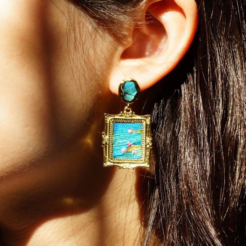 May include: A gold-toned earring featuring a rectangular frame with an ornate border. The center displays a textured blue surface with small pink floral accents. A matching circular blue stud sits above the rectangular drop component.