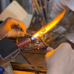 Pode incluir: Uma foto em close-up de uma joia sendo feita com um ma&ccedil;arico. A pe&ccedil;a &eacute; dourada com um detalhe azul. A chama &eacute; laranja brilhante e amarela, iluminando as m&atilde;os do artes&atilde;o no trabalho.