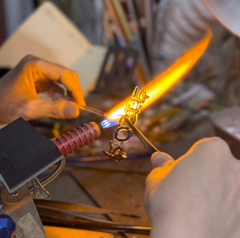 Pode incluir: Uma foto em close-up de uma joia sendo feita com um ma&ccedil;arico. A pe&ccedil;a &eacute; dourada com um detalhe azul. A chama &eacute; laranja brilhante e amarela, iluminando as m&atilde;os do artes&atilde;o no trabalho.