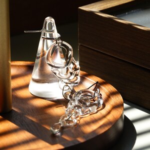 Pode incluir: Brincos de vidro transparente com formas irregulares, apresentados num suporte c&oacute;nico transparente e numa bandeja de madeira. A luz solar cria reflexos. Uma caixa de madeira &eacute; vis&iacute;vel no fundo. Joias modernas.