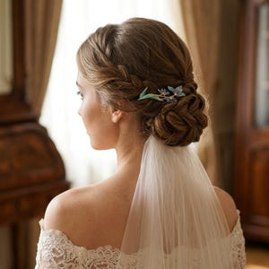 May include: A hair accessory featuring a delicate blue and green floral design secured in a braided updo hairstyle. The accessory is positioned on the side of the head, complementing a white lace garment and a sheer white veil draped down the back.