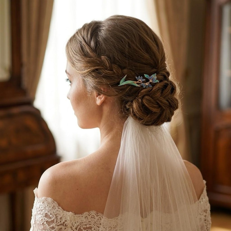 May include: A hair accessory featuring a delicate blue and green floral design secured in a braided updo hairstyle. The accessory is positioned on the side of the head, complementing a white lace garment and a sheer white veil draped down the back.