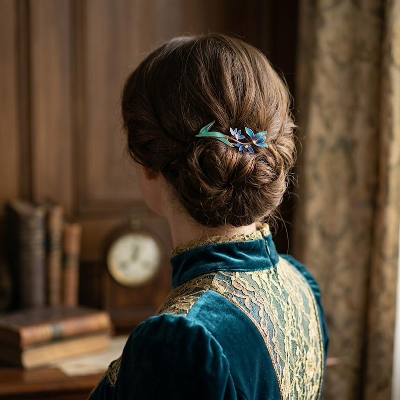 May include: A hair accessory featuring a delicate floral design with blue and green enamel petals and small white accents, secured in a brown hair bun. The piece is worn with a dark teal velvet garment featuring intricate gold lace detailing.