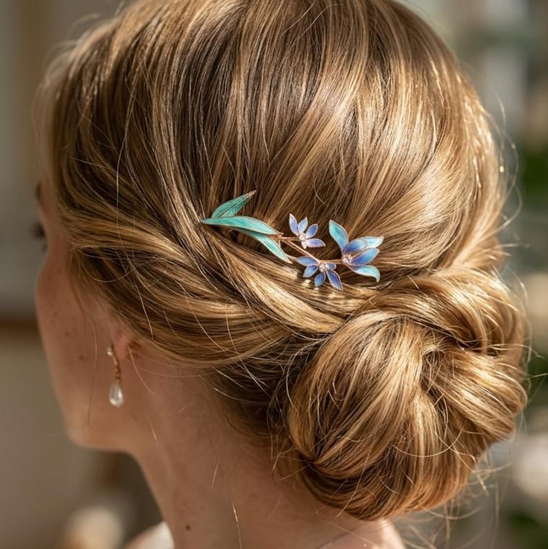 May include: A hair accessory featuring a metallic branch design with delicate blue and green enamelled leaves and small white floral accents. The piece is secured into a blonde hair bun, showcasing a slender stem and multiple petal-like elements.