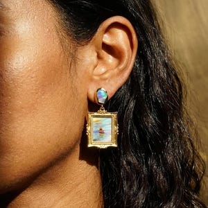 May include: A gold-toned drop earring featuring a small, iridescent oval opal at the top, connected to a rectangular, ornate gold-toned frame. The frame contains a miniature landscape painting depicting a sunset over water with soft brushstrokes.