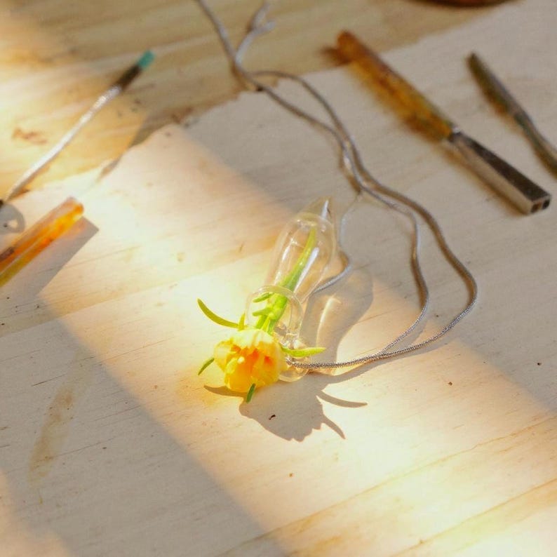Pu&ograve; includere: Un ciondolo in vetro trasparente a forma di piccola fiala contenente un singolo fiore giallo, fissato a una sottile catenina in metallo argentato. Il ciondolo poggia su una superficie chiara accanto a un utensile in metallo e un bastoncino di legno.