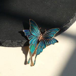 May include: A detailed butterfly brooch with teal and blue enamel wings and a gold-tone frame. The brooch has a delicate design with intricate details, and the wings are spread out. The butterfly is a decorative accessory.