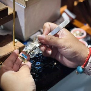 May include: A close-up of jewelry being crafted. A person is using tweezers to work on a small, gold-colored pendant with a teardrop shape. The pendant has a blue accent. The background is blurred, showing a workbench.