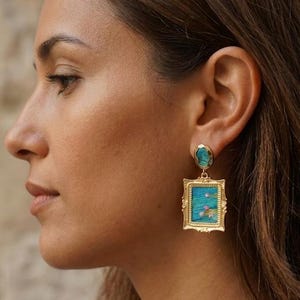 May include: A gold-toned earring featuring a small, irregular turquoise-colored stone at the earlobe, connected to a rectangular, ornate gold-toned frame pendant that displays a miniature blue landscape painting with small pink floral accents.
