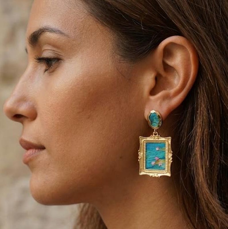 May include: A gold-toned earring featuring a small, irregular turquoise-colored stone at the earlobe, connected to a rectangular, ornate gold-toned frame pendant that displays a miniature blue landscape painting with small pink floral accents.