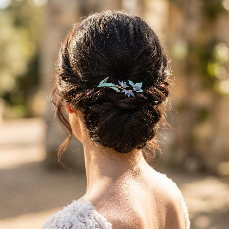 May include: A hair accessory featuring a delicate floral design with light blue and green enamel petals and leaves. The piece is secured into dark brown hair styled in an intricate braided updo, positioned horizontally against the back of the head.