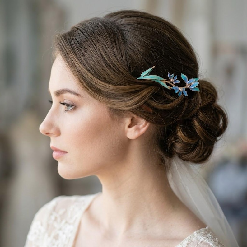 May include: A hair comb featuring a delicate metal branch design with small, vibrant blue and teal enamelled leaves and flowers. The accessory is secured into a styled brown updo, showcasing a metallic stem that curves along the side of the hair.