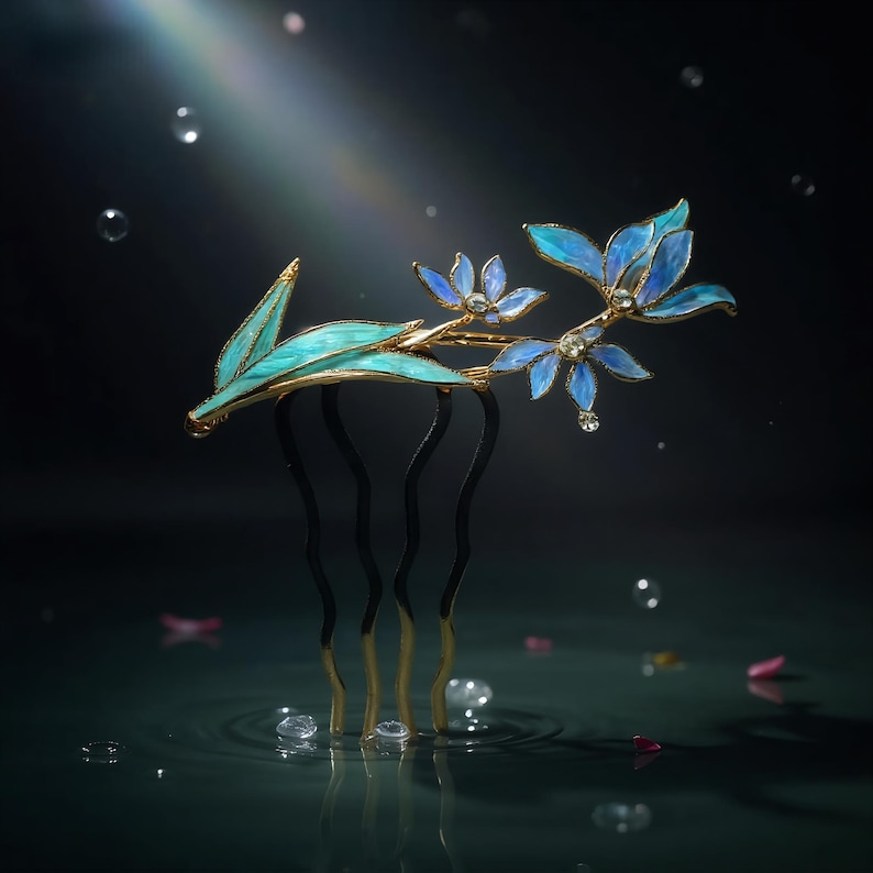 May include: A gold-toned hair comb featuring delicate, translucent blue and teal enamel flower petals and leaves. The comb has four wavy prongs and is accented with small, clear crystals that catch the light, creating a floral design.