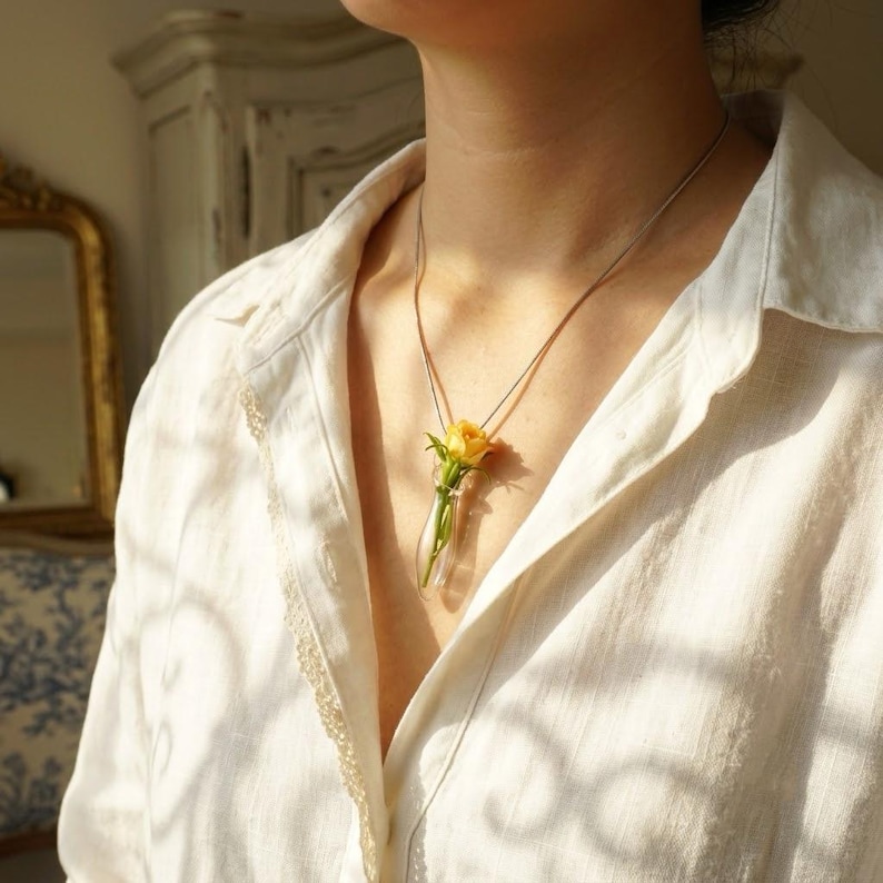 Pu&ograve; includere: Una collana con ciondolo a forma di fiala in vetro trasparente contenente un piccolo bocciolo di rosa gialla. Il ciondolo pende da una sottile catenina color argento, appoggiato su una camicia bianca con colletto. Il vetro &egrave; trasparente.