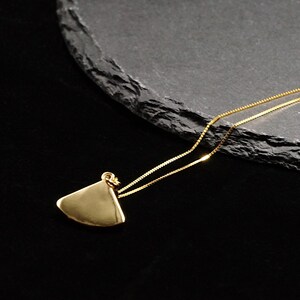 May include: A gold-colored pendant necklace with a triangular shape. The pendant hangs from a delicate gold chain. The necklace is displayed on a dark, textured surface against a black background. The pendant has a smooth, polished finish.