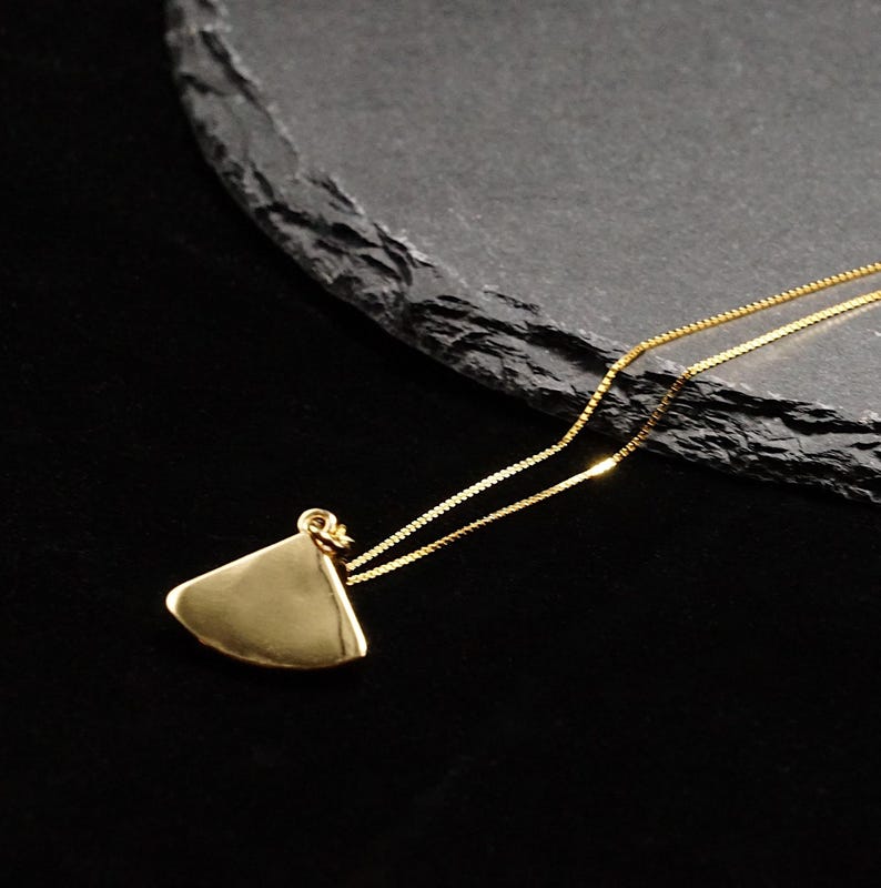 May include: A gold-colored pendant necklace with a triangular shape. The pendant hangs from a delicate gold chain. The necklace is displayed on a dark, textured surface against a black background. The pendant has a smooth, polished finish.