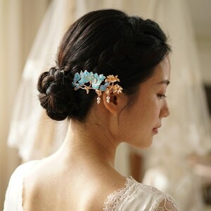 Pode incluir: Um pente de cabelo decorativo com motivos florais de metal em tons de azul turquesa e dourado. O design inclui folhas e p&eacute;talas delicadas com pequenas contas brancas semelhantes a p&eacute;rolas penduradas na base das flores de metal.
