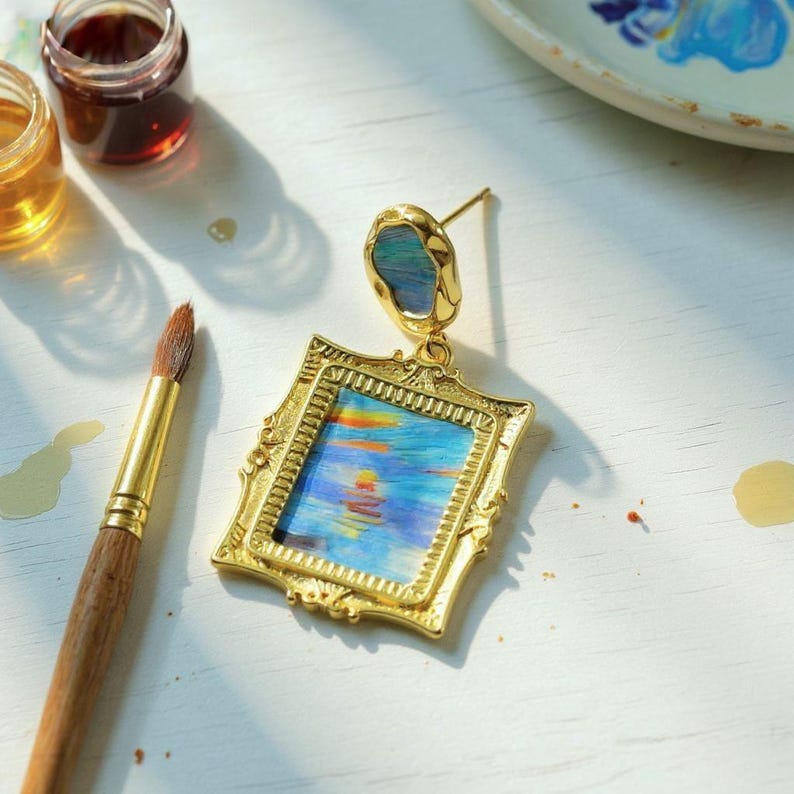 May include: A gold-toned earring featuring a rectangular frame pendant with an ornate border. The center of the frame displays a miniature painting of a sunset over water in shades of blue, orange, and yellow. A small, irregular blue stone sits above.