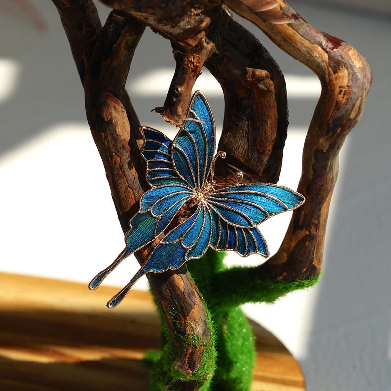 May include: A detailed, blue butterfly brooch with gold-coloured trim. The butterfly is perched on a natural, brown branch with green moss. The butterfly's wings have a gradient of blue hues.