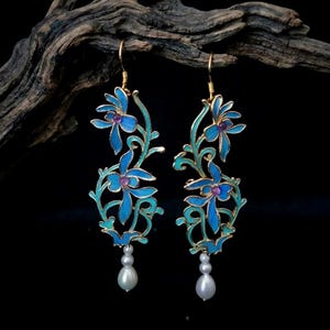 May include: A pair of floral earrings with a gold-tone metal frame. The earrings feature blue enamel flowers with purple centers and turquoise stems. Each earring has a white teardrop-shaped pearl pendant. The earrings are displayed against a dark background.