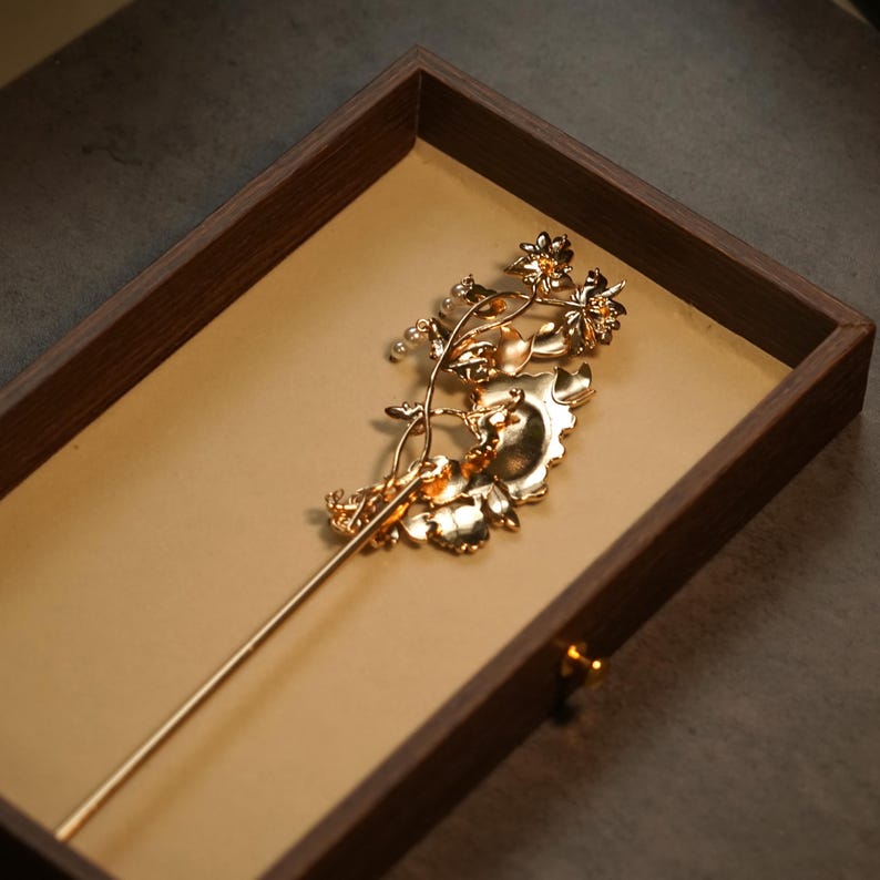 Pode incluir: Um gancho de cabelo de metal em tom dourado com um design floral intrincado, folhas delicadas e pequenos detalhes esf&eacute;ricos brancos. A cabe&ccedil;a decorativa do gancho tem a forma de um cacho de flores e folhagem, estendendo-se numa haste fina.