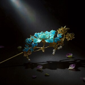 Pode incluir: Um gancho de cabelo dourado com um design floral intrincado com p&eacute;talas e folhas em esmalte azul vibrante. A pe&ccedil;a decorativa inclui pequenas p&eacute;rolas brancas pendentes presas aos caules, criando um acess&oacute;rio de cabelo delicado e detalhado.