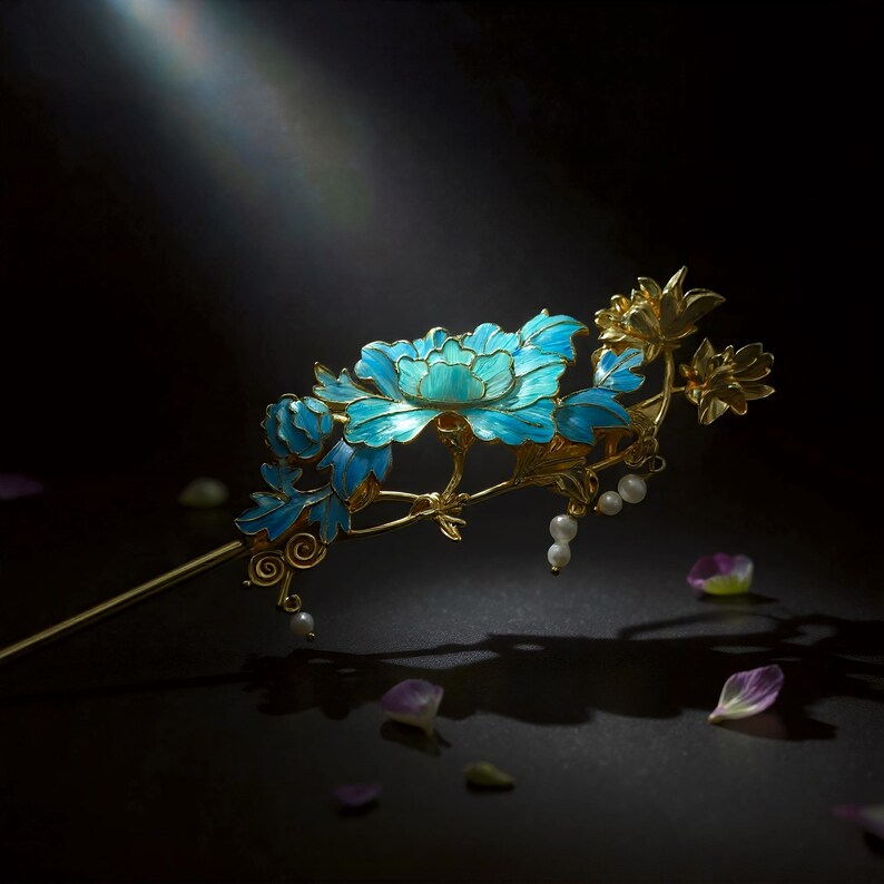 Pode incluir: Um gancho de cabelo dourado com um design floral intrincado com p&eacute;talas e folhas em esmalte azul vibrante. A pe&ccedil;a decorativa inclui pequenas p&eacute;rolas brancas pendentes presas aos caules, criando um acess&oacute;rio de cabelo delicado e detalhado.