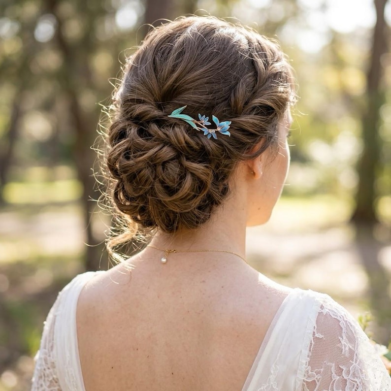 May include: A hair accessory featuring a delicate gold-tone metal stem with light blue enamel flower petals and green leaves, secured into a brown braided updo hairstyle. The accessory is positioned on the side of the hair, adding a floral accent.