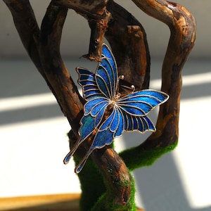 May include: A blue butterfly brooch with gold-coloured edging. The butterfly is displayed on a brown, textured branch with green moss. The wings feature a gradient of blue shades.